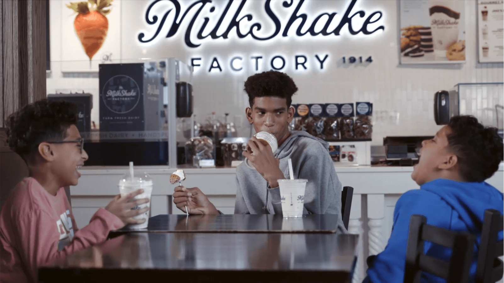 three boys sitting at table inside milkshake factory store drinking milkshakes